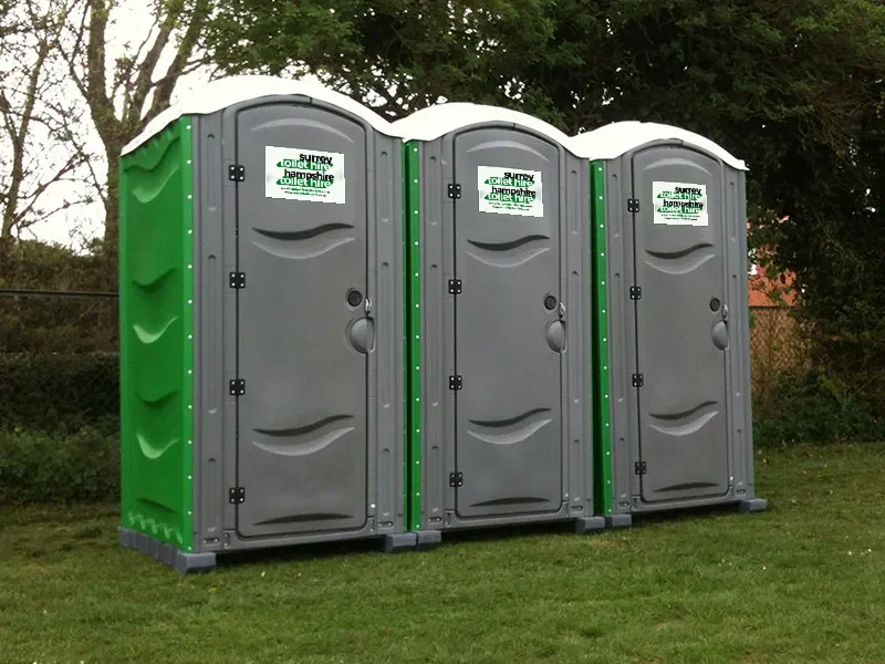 Three of our portable toilets standing on grass by a hedge, one green and two grey