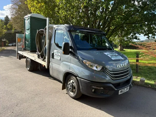 Our grey flatbed truck carrying a green portable toilet, with Surrey and Hampshire Toilet Hire stickers on the bonnet and door