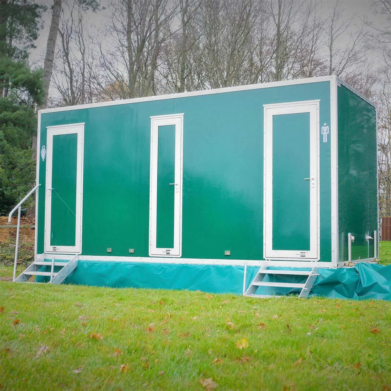 A green luxury toilet trailer with steps at each door, set up on a lawn beside trees
