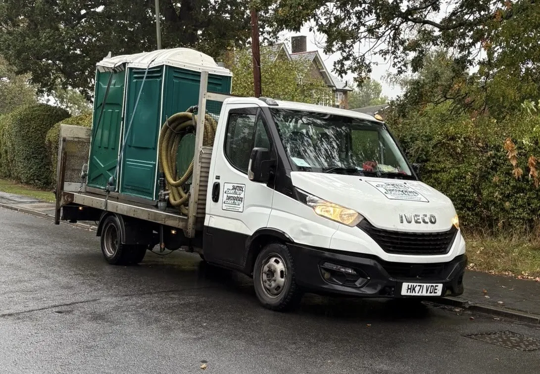Our white pump truck with two green portable toilets and its hose reel on the back, parked in a street